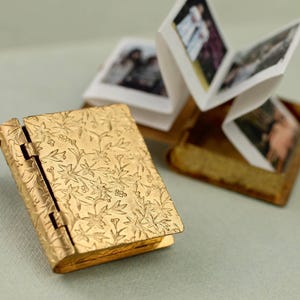 May include: A gold-toned metal locket shaped like a miniature book with an intricate floral embossed cover. The locket is attached to a delicate chain and is shown open, revealing a concertina-style paper insert featuring small printed photographs.