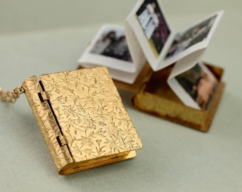 Può includere: Un medaglione in metallo dorato a forma di libro in miniatura con una copertina finemente goffrata a motivi floreali. Il medaglione è fissato a una catenina sottile ed è mostrato aperto, rivelando un inserto in carta a fisarmonica con foto.