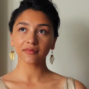 May include: A woman wearing gold-colored earrings. The earrings have a fan-like design with multiple vertical bars. The woman has dark hair and is looking upwards. The earrings are a stylish accessory.