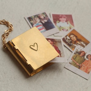 Puede incluir: Un relicario de metal dorado con forma de pequeño libro con un corazón grabado en la portada, unido a una delicada cadena dorada. El relicario está parcialmente abierto, revelando una pequeña fotografía cuadrada en su interior.