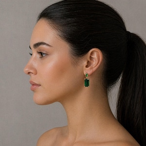 May include: Gold-coloured earrings featuring rectangular emerald-green gemstones. The earrings have a classic design, with a smaller stone at the top and a larger, elongated stone below. The earrings are worn on a person's ear.