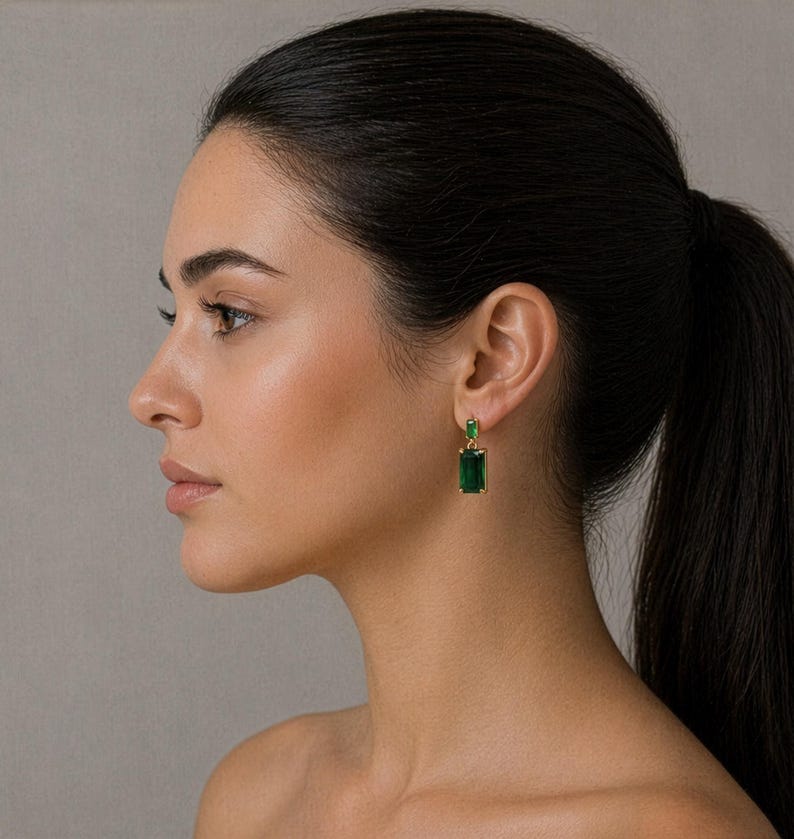 May include: Gold-coloured earrings featuring rectangular emerald-green gemstones. The earrings have a classic design, with a smaller stone at the top and a larger, elongated stone below. The earrings are worn on a person's ear.