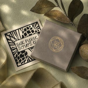 May include: A gray square box and a card with the text "Silk Purse Sows Ear" in black and gold. The box has a gold logo with the same text. The background is a light green with a ribbon and leaves.
