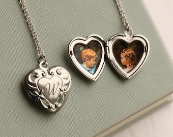 May include: Silver heart-shaped locket necklace with a chain. One locket is closed, displaying the letter "W", while the other is open, revealing two small portraits. The locket is on a light green surface.