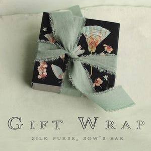 May include: A small black gift box with a floral design, tied with a sage green ribbon. The box features illustrations of butterflies and flowers. The text "GIFT WRAP" is displayed below the box.