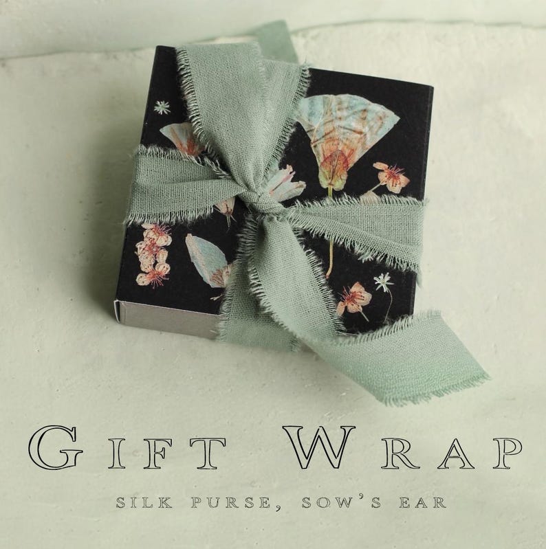 May include: A small black gift box with a floral design, tied with a sage green ribbon. The box features illustrations of butterflies and flowers. The text "GIFT WRAP" is displayed below the box.