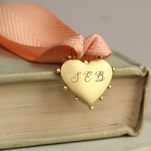 May include: A gold heart-shaped charm with the initials "SEB" engraved on it. The charm is attached to a peach-colored ribbon.