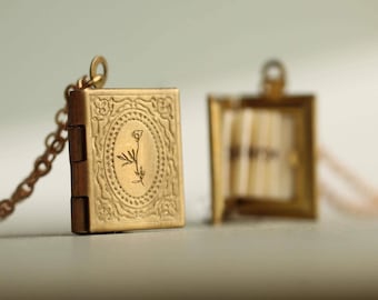 May include: A gold-toned metal locket shaped like a miniature book with an embossed floral design on the cover. The locket is attached to a delicate chain. A second, identical locket is shown open, revealing small, cream-coloured paper pages inside.