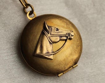 Könnte beinhalten: Ein rundes, messingfarbenes Metallmedaillon mit einem erhabenen Relief eines Pferdekopfes im Profil, komplett mit Zaumzeug. Das Medaillon ist oben an einem passenden Metallkettenglied befestigt und hat einen sichtbaren Scharniermechanismus unten rechts.
