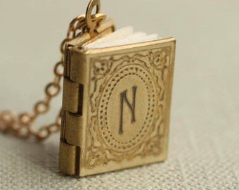 May include: A miniature book-shaped pendant made of brass-toned metal, featuring an embossed decorative border and the letter N centered on the front cover. The pendant is attached to a matching metal chain and has visible white paper pages.