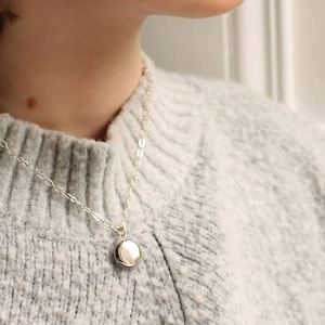 May include: A silver-toned necklace featuring a delicate chain and a small, smooth, circular pendant. The pendant has a polished, reflective surface that catches the light, suspended from a simple link chain resting against a textured grey sweater.