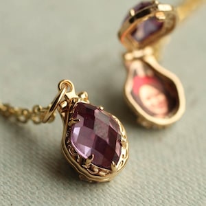 May include: A gold-toned locket necklace featuring a faceted, pear-shaped purple gemstone set in an ornate metal frame. The locket is shown in two positions: one closed, displaying the purple stone, and one open, revealing a small, blurry portrait inside.