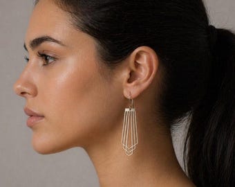 Puede incluir: Un par de pendientes geométricos plateados de estilo Art Déco. Los pendientes tienen una parte superior rectangular con líneas verticales que se abren en abanico en una serie de formas geométricas escalonadas. Los pendientes cuelgan de un gancho plateado.