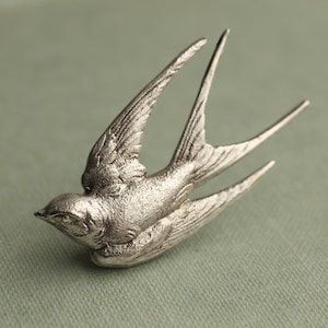 Pode incluir: Um broche de metal prateado em forma de andorinha em voo. A ave é representada com as asas estendidas para cima e uma cauda bifurcada. A superfície do metal tem um acabamento fosco e texturizado que destaca os detalhes das penas.