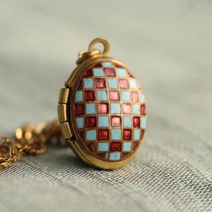 May include: An oval gold locket pendant featuring a checkered pattern of alternating light blue and deep red enamel squares. The locket has a hinged side and is attached to a gold chain, showcasing a metallic frame around the colorful grid.