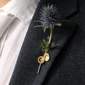 Peut inclure: Une boutonnière composée d'une fleur de panicaut bleu avec des feuilles vertes et d'un médaillon en métal doré attaché à la tige. Le médaillon est ouvert, révélant deux petits portraits circulaires à l'intérieur, épinglé sur un revers de blazer gris anthracite texturé.