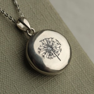 Op de afbeelding: Een zilverkleurige hangerketting met een ronde medaillon met een gedetailleerd zwart paardenbloemontwerp. De hanger hangt aan een fijne zilveren ketting. Het medaillon is glad en gepolijst en reflecteert licht.