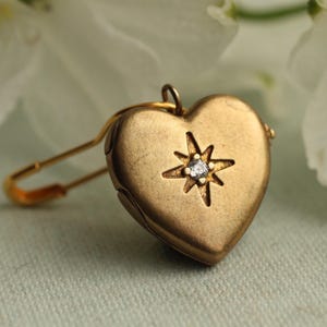 May include: A gold-toned heart-shaped locket attached to a safety pin. The locket features a matte finish and a central starburst engraving with a small, clear, round-cut stone set in the middle. The piece has a small loop at the top for attachment.