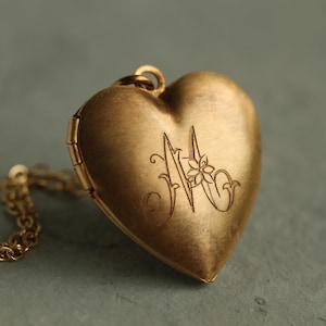 Puede incluir: Un colgante de relicario en forma de corazón de oro con acabado mate, que presenta una letra M grabada con un pequeño detalle floral integrado en el diseño. El relicario está unido a un collar de cadena de eslabones de oro a juego con una bisagra visible.