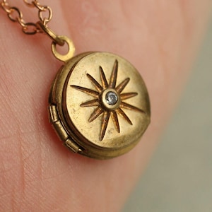 May include: A round gold-toned metal locket pendant suspended from a matching chain. The front of the locket features an engraved starburst pattern with a single small clear stone set in the centre. The metal has a matte, slightly weathered finish.