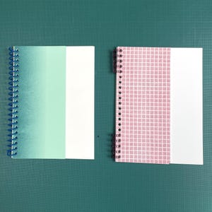 Peut inclure: Deux cahiers à spirales. L'un a une couverture dégradée du turquoise au blanc, l'autre un motif quadrillé rose. Les deux ont des pages de papier blanc. Les cahiers sont ouverts, montrant les pages intérieures.