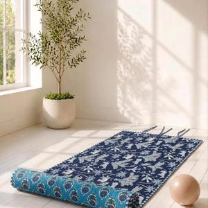 May include: A rectangular quilted mat featuring a dark blue surface with a white botanical and animal print pattern. One end is rolled up, revealing a bright turquoise underside with a repeating dark blue and green floral motif and stitched edges.