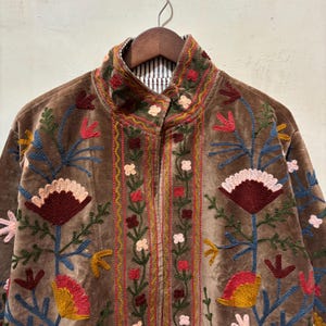 May include: A brown velvet jacket adorned with vibrant floral embroidery. The jacket has a high collar and a striped lining at the neck. The embroidery features red, pink, yellow, and blue flowers and leaves, creating a colourful design.