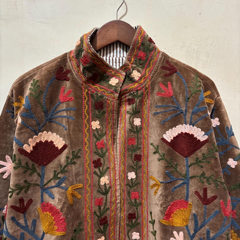 May include: A brown velvet jacket adorned with vibrant floral embroidery. The jacket has a high collar and a striped lining at the neck. The embroidery features red, pink, yellow, and blue flowers and leaves, creating a colourful design.