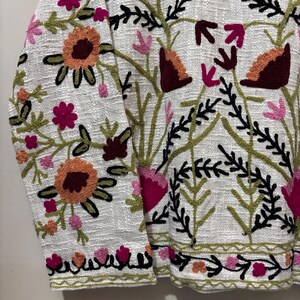 May include: White jacket with colorful floral embroidery. The embroidery features orange, pink, and burgundy flowers with green leaves and black outlines. The jacket has a textured fabric and a detailed floral pattern.