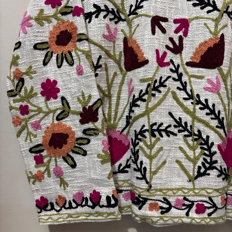 May include: White jacket with colorful floral embroidery. The embroidery features orange, pink, and burgundy flowers with green leaves and black outlines. The jacket has a textured fabric and a detailed floral pattern.