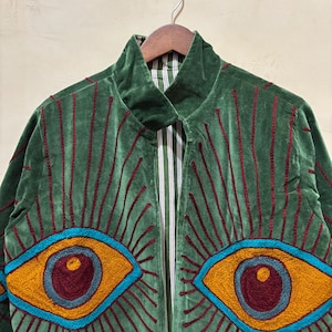 May include: Green velvet jacket with embroidered eye design. The jacket features a high collar and a striped lining. The eyes are outlined in blue, with yellow irises and maroon pupils, and red embroidered rays.