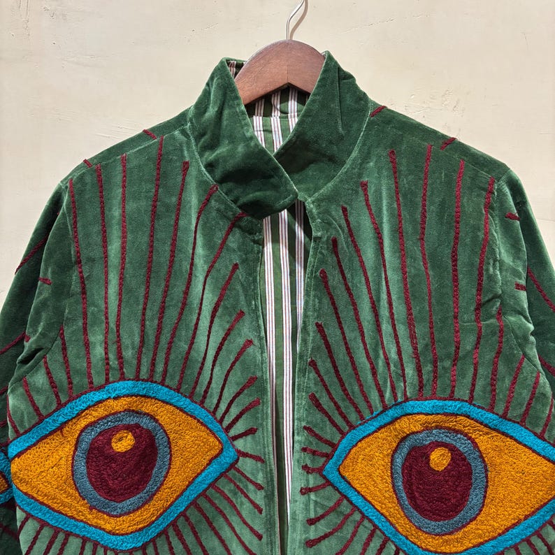 May include: Green velvet jacket with embroidered eye design. The jacket features a high collar and a striped lining. The eyes are outlined in blue, with yellow irises and maroon pupils, and red embroidered rays.