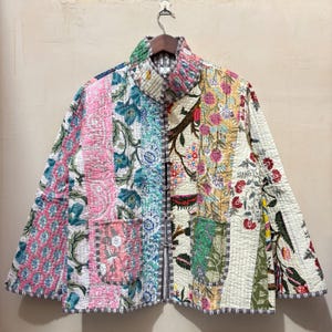 May include: A quilted jacket with a patchwork design. The jacket features a variety of floral patterns in shades of pink, blue, green, and white. It has a stand-up collar, two pockets, and a button-up front. The jacket is hanging on a wooden hanger.