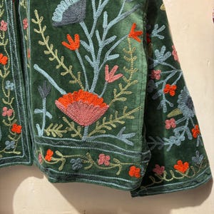May include: A dark green velvet jacket with intricate floral embroidery. The embroidery features orange, pink, and blue flowers and leaves. The jacket has a detailed border design and is a unique piece of apparel.