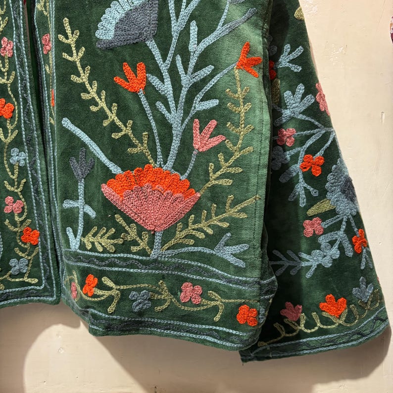 May include: A dark green velvet jacket with intricate floral embroidery. The embroidery features orange, pink, and blue flowers and leaves. The jacket has a detailed border design and is a unique piece of apparel.