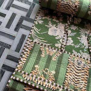 May include: A quilted green fabric roll featuring a repeating pattern of tigers and floral motifs. The textile has a striped green background and is finished with a thin, red and white striped fabric trim along the edges and tie closures.