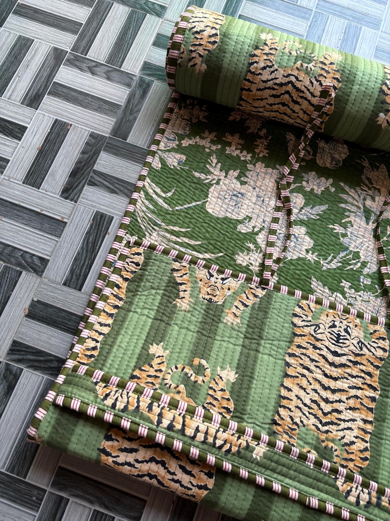 May include: A quilted green fabric roll featuring a repeating pattern of tigers and floral motifs. The textile has a striped green background and is finished with a thin, red and white striped fabric trim along the edges and tie closures.