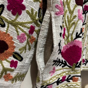 May include: Close-up of a light-colored jacket with intricate floral embroidery. The jacket features a pocket and is adorned with colorful embroidered flowers in shades of pink, orange, and green, creating a textured, artistic design.