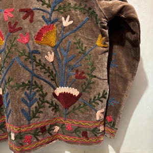 May include: A brown velvet top with detailed floral embroidery. The embroidery uses red, yellow, pink, blue, and green. The design includes stylized flowers and leaves, with a detailed border along the bottom.