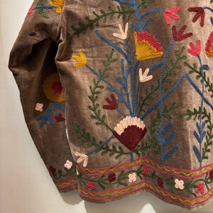 May include: A brown velvet jacket with colourful floral embroidery. The embroidery features yellow, red, pink, and blue flowers and green leaves. The jacket has long sleeves and a detailed embroidered border along the bottom.