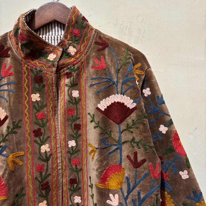 May include: A brown velvet jacket with colourful floral embroidery. The jacket has a high collar and a striped lining. The embroidery features red, pink, yellow, and blue flowers and leaves, creating a detailed and artistic design.