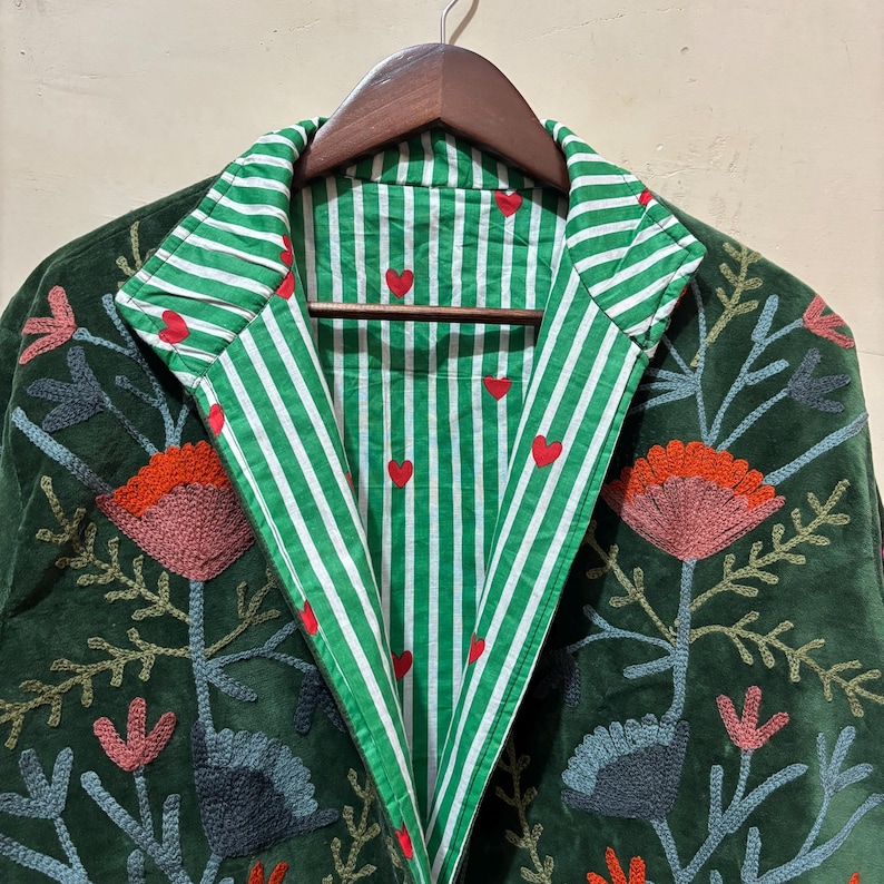 May include: A dark green velvet jacket with floral embroidery in shades of orange, pink, and gray. The interior lining features green and white stripes with red heart accents. The jacket is hanging on a wooden hanger.