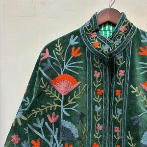 May include: A dark green velvet jacket with colorful floral embroidery. The jacket features a high collar and a striped green and white lining. The embroidery includes orange, pink, and blue flowers and leaves, creating a detailed and artistic design.