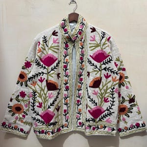 May include: A white jacket with vibrant floral embroidery. The jacket features large pink flowers, orange accents, and green leaves, all outlined in black thread. The collar and front edges are also adorned with floral patterns, creating a bohemian aesthetic.