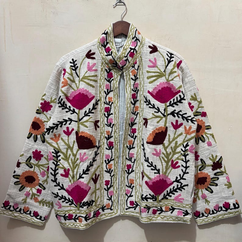 May include: A white jacket with vibrant floral embroidery. The jacket features large pink flowers, orange accents, and green leaves, all outlined in black thread. The collar and front edges are also adorned with floral patterns, creating a bohemian aesthetic.