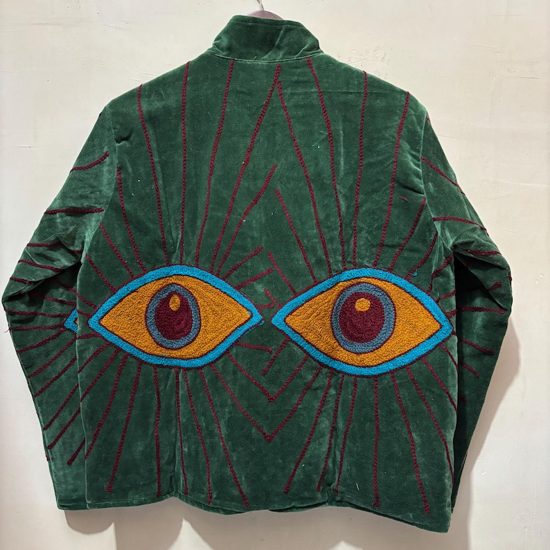 May include: A green velvet jacket with embroidered eye design on the back. The eyes are outlined in blue with orange and maroon details. Red lines radiate from the eyes, and the jacket has a high collar.