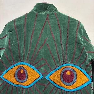May include: Green velvet jacket with embroidered details. The back features a design of radiating lines and two large eyes in shades of yellow, blue, and burgundy. The jacket has a high collar and is hanging on a hanger.