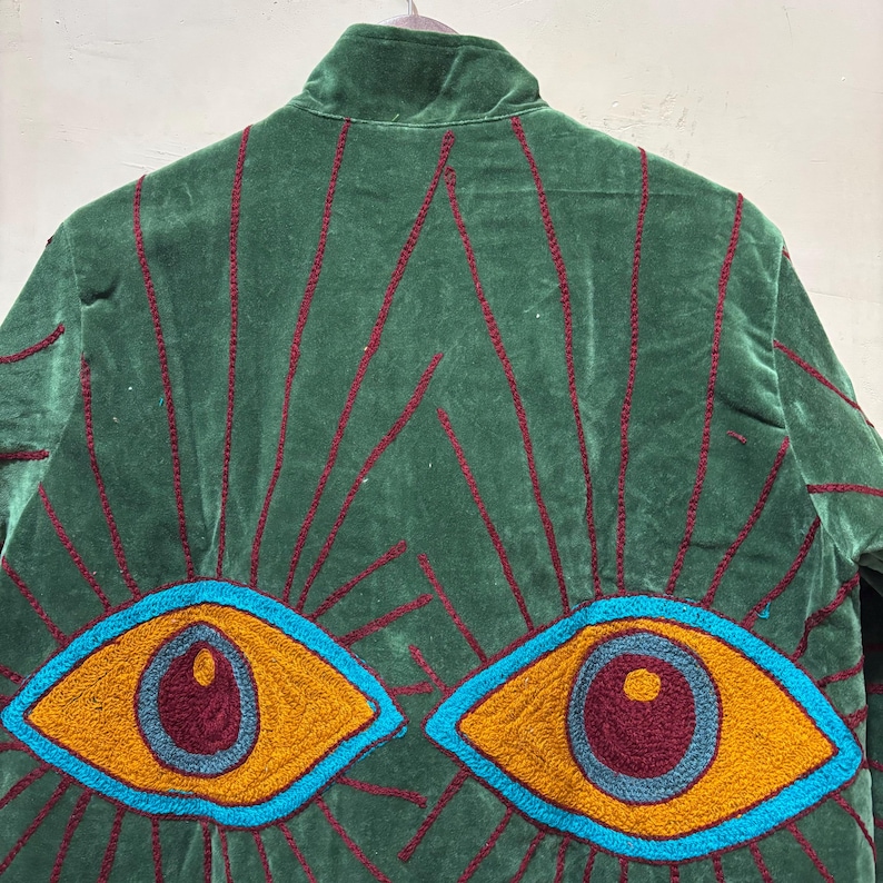 May include: Green velvet jacket with embroidered details. The back features a design of radiating lines and two large eyes in shades of yellow, blue, and burgundy. The jacket has a high collar and is hanging on a hanger.