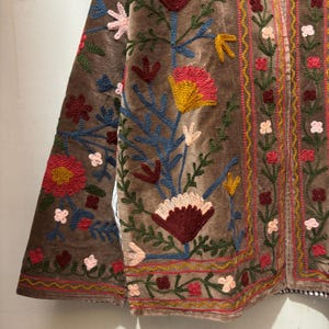 May include: Brown velvet jacket with vibrant floral embroidery. The jacket showcases red, yellow, blue, and pink flowers with green foliage. The detailed embroidery adorns the front and sleeves of the garment.
