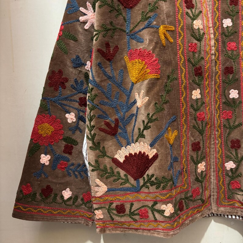 May include: Brown velvet jacket with vibrant floral embroidery. The jacket showcases red, yellow, blue, and pink flowers with green foliage. The detailed embroidery adorns the front and sleeves of the garment.